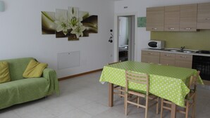 Un salon avec un canapé vert, une table à manger recouverte d’une nappe verte et un coin cuisine doté de meubles en bois et d’un four à micro-ondes.