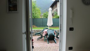Une porte ouverte donne sur un patio extérieur agrémenté de chaises vertes, d’une table et d’un parasol blanc.