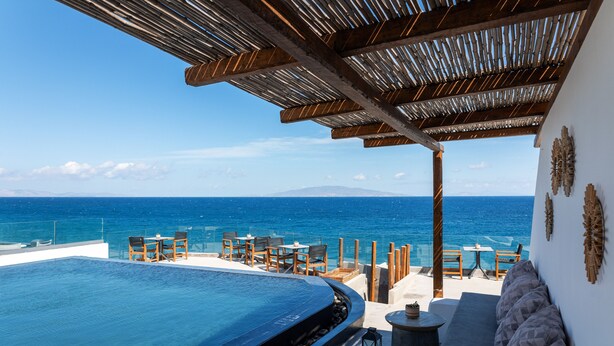A infinity pool with a view of the ocean, wooden furniture, and a covered seating area. - Almyra Oia Suites Adults Only  (Santorini)