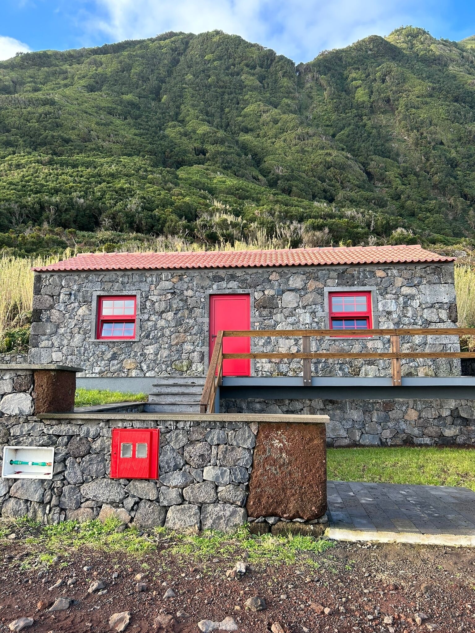A stone building with a red roof and two red doors, set against a backdrop of lush green hills.