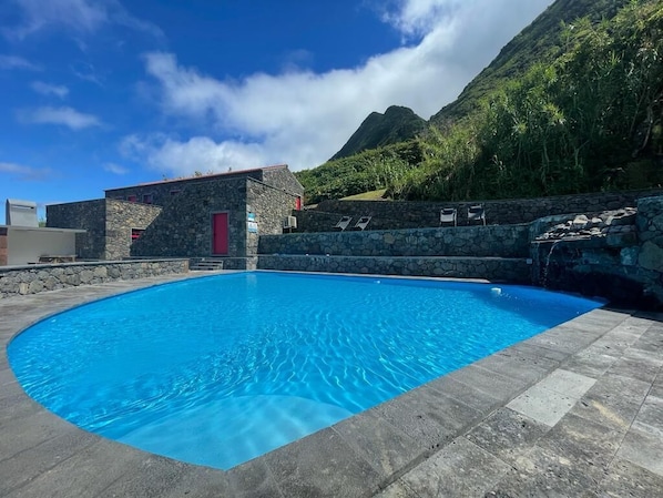 A swimming pool with a stone wall and a red door, surrounded by a mountainous landscape.