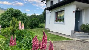 En vit byggnad med ett mörkt tak, en liten trädgård med rosa blommor och en klar himmel.