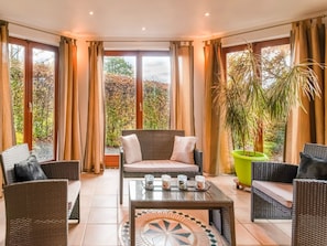Une pièce meublée de mobilier en osier, avec une table en verre décorée de bougies et de grandes fenêtres ornées de rideaux.