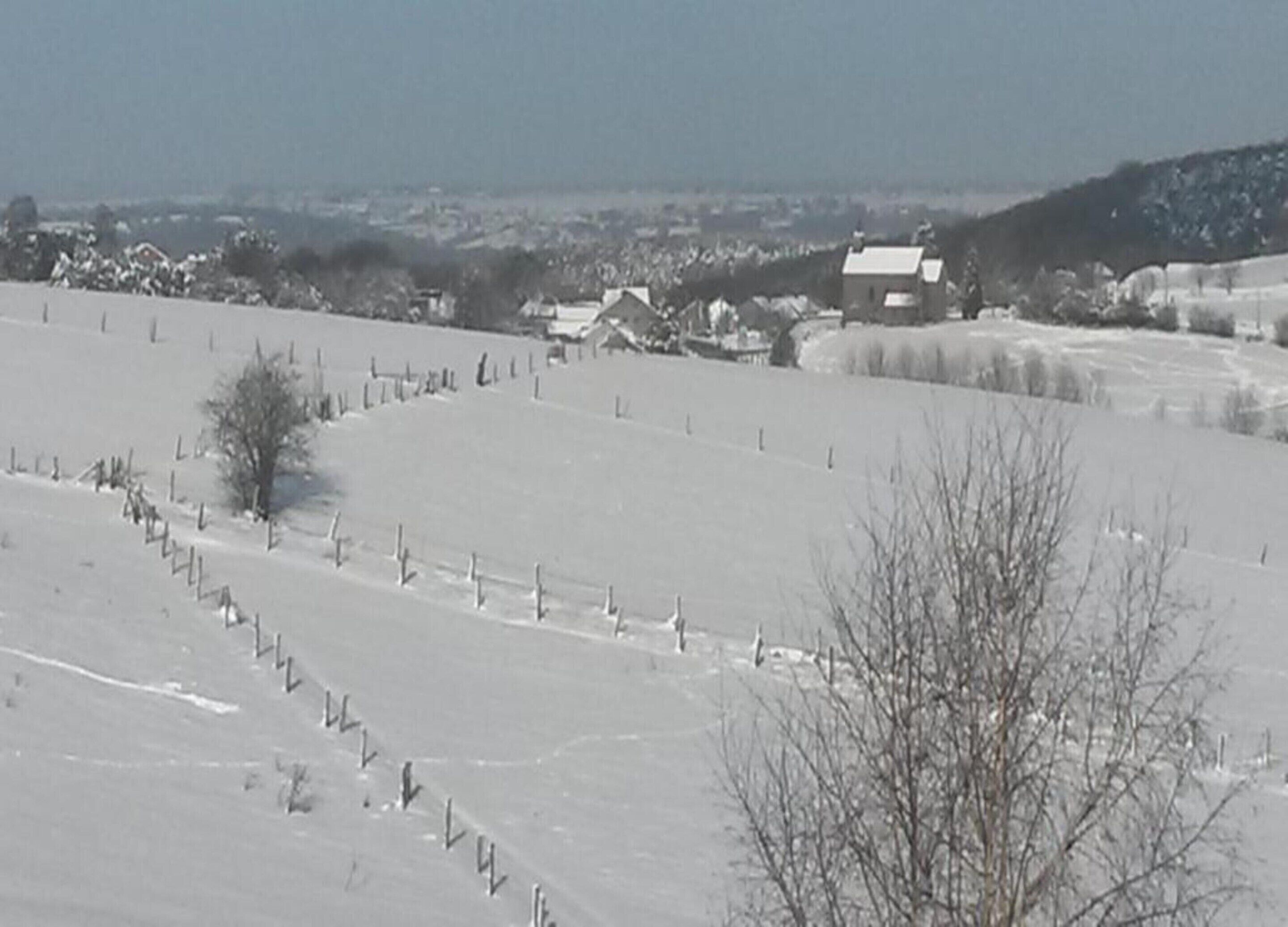 Un paisaje nevado con una zona cercada, algunos edificios y una colina al fondo.