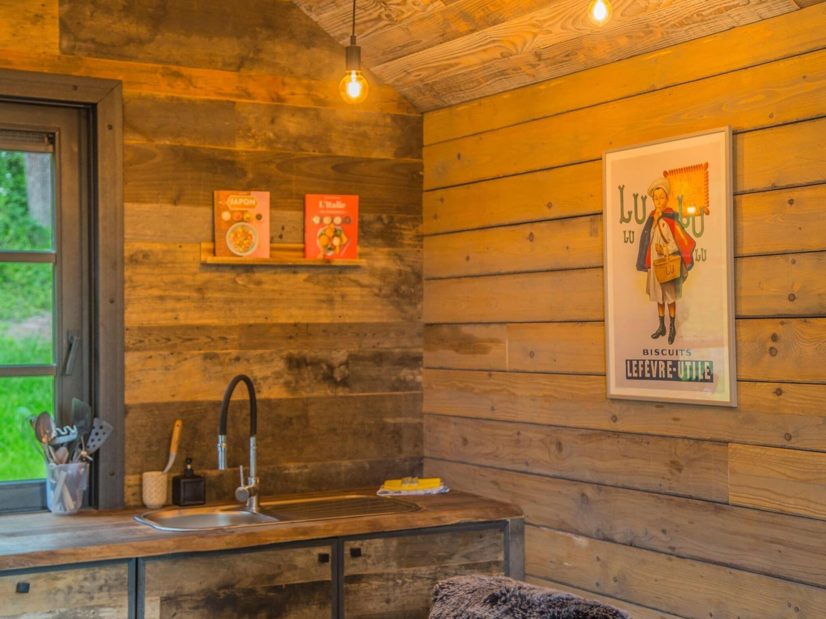 Een keuken met houten wanden, een spoelbak en een hangende lamp. Twee ingekaderde posters hangen aan de muur.