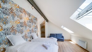 Ett sovrum med ett sluttande tak, ett stort fönster, en blå soffa och en vägg med mönster.