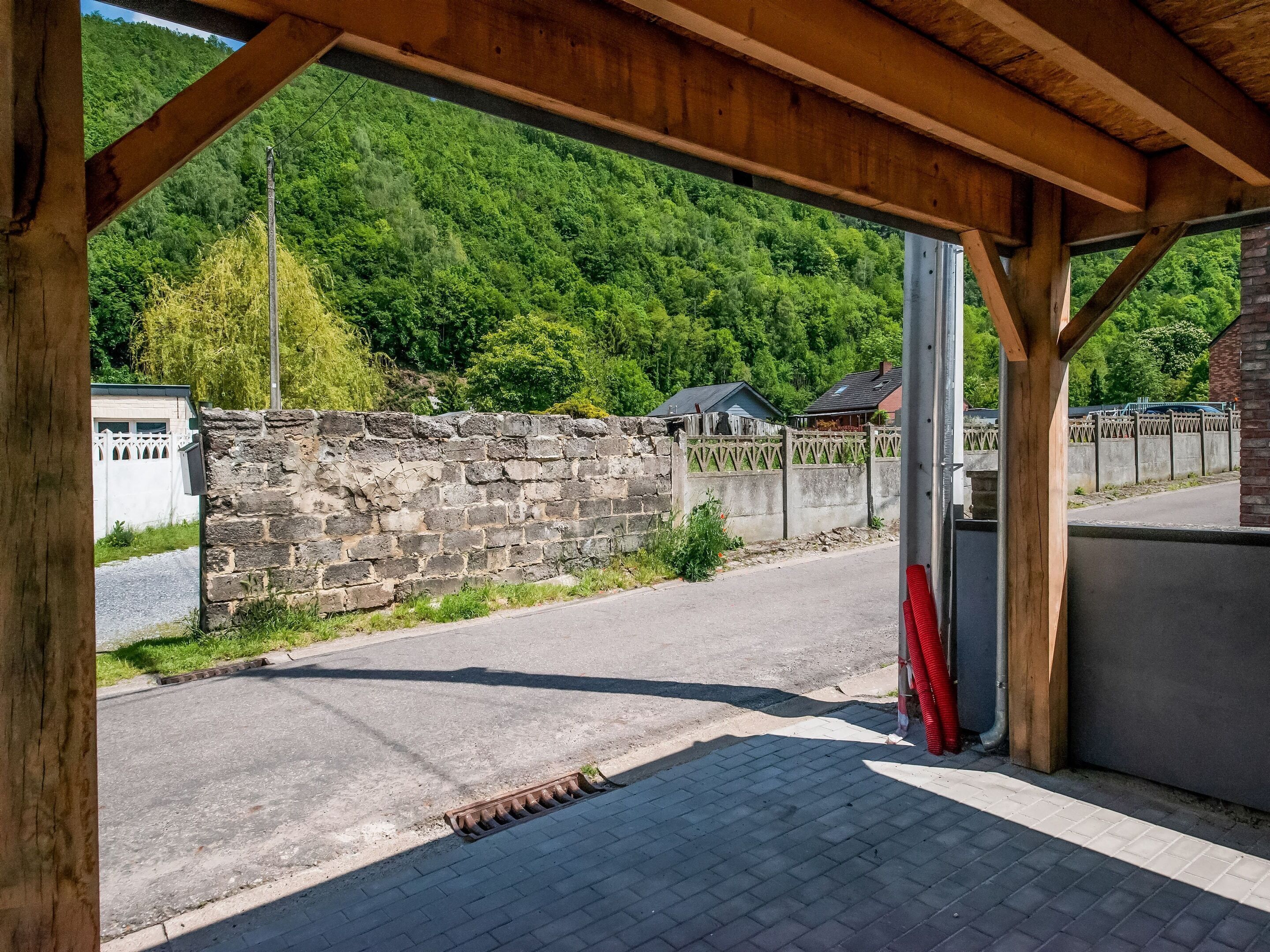 A covered outdoor area with a stone wall, a paved road, and a view of a forested hillside.