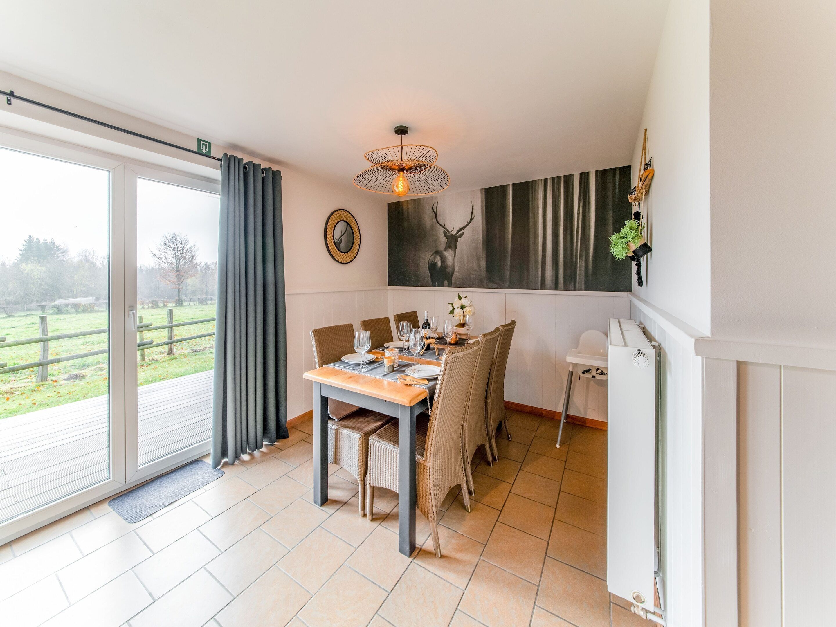 A dining area with a table set for four, a large window with curtains, a wall-mounted mirror, and a deer painting.