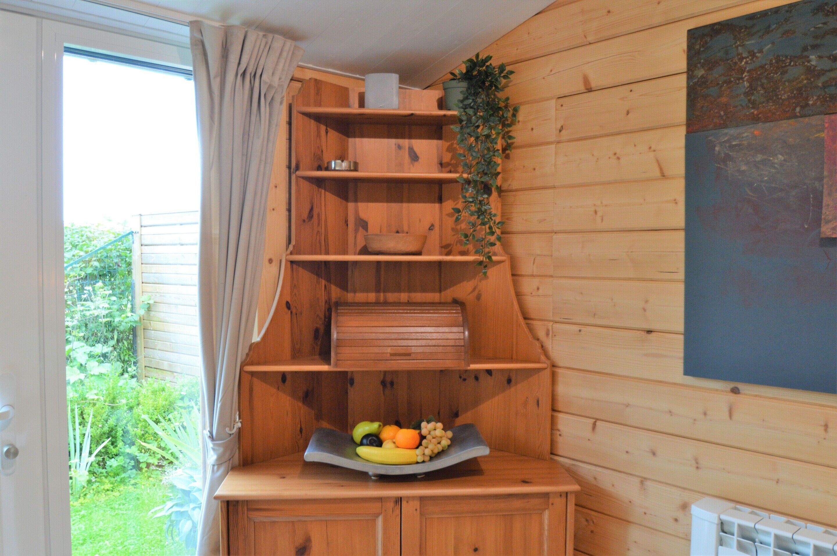 Un intérieur en bois avec un panier de fruits posé sur une étagère, une plante suspendue et une fenêtre avec des rideaux.