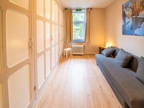 A room with a wooden floor, a grey sofa, a radiator, a small stool, and a window with curtains. - Holiday Home in Falaen by Chateau Montaigle (Onhaye)