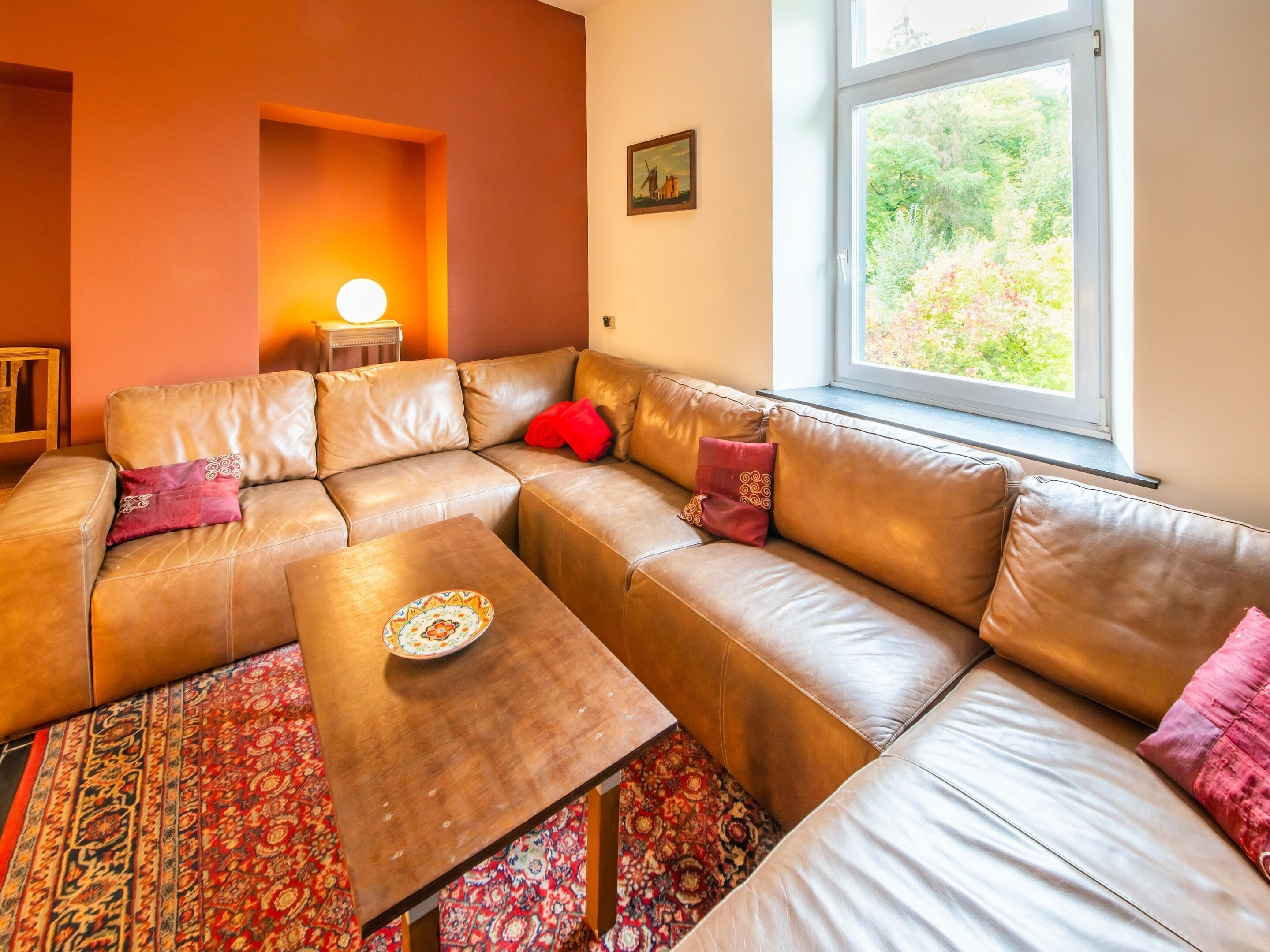 Ein modernes Wohnzimmer mit einer Eckcouch, einem Couchtisch, einem gemusterten Teppich, einem Fenster mit Blick auf Grünflächen und einer Wandlampe.