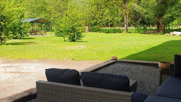 Eine Terrassenfläche mit Korbmöbeln und Kissen, ein kleiner Garten mit Bank und Baum sowie freie Sicht auf die umliegende Natur.