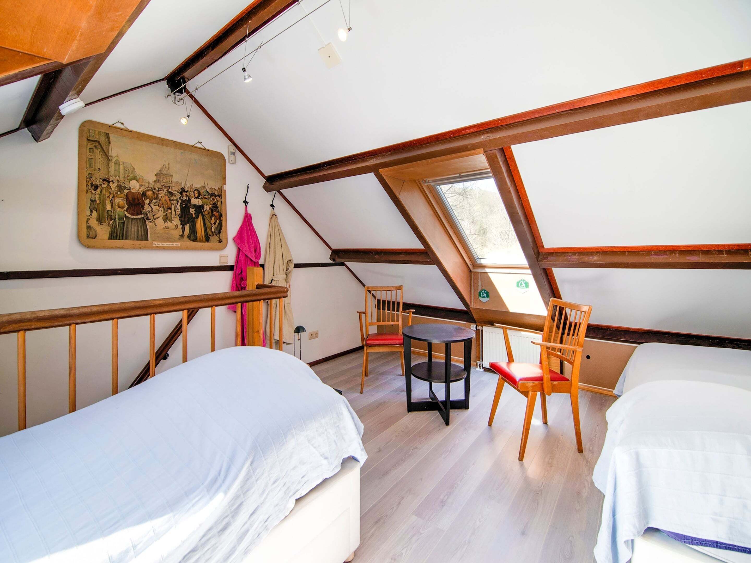 Een kamer met twee bedden, een kleine tafel en een stoel. Er hangt een schilderij aan de muur en een raam zorgt voor natuurlijk licht.