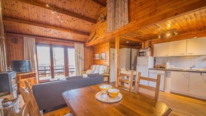 L’intérieur chaleureux d’un chalet avec une table à manger en bois, un canapé, un coin cuisine et un téléviseur dans le salon.