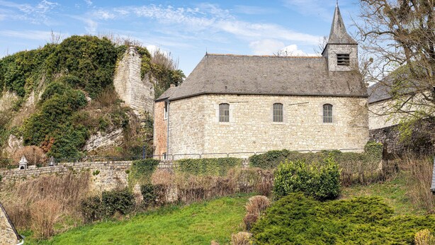 Un edificio de piedra con un campanario, rodeado de vegetación y un muro de piedra.