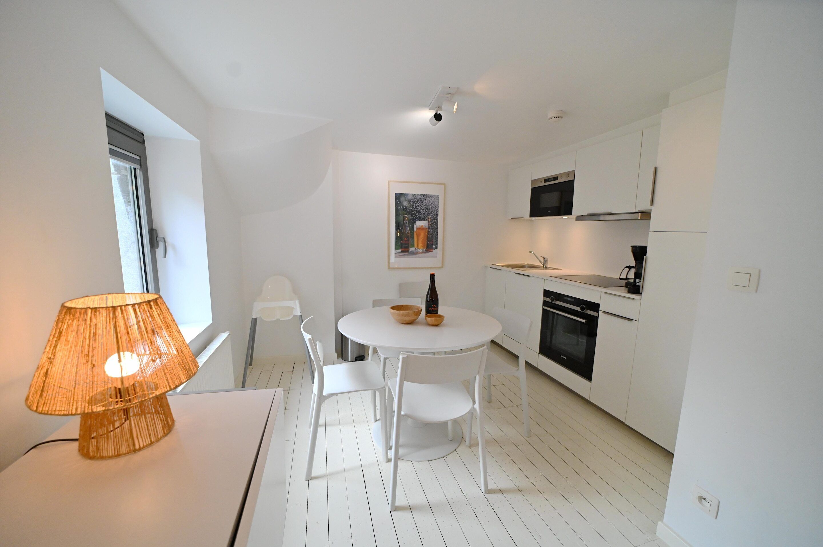 A modern kitchen with white cabinets, a dining table with chairs, a lamp, and a painting on the wall.