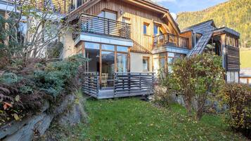 A modern wooden house with large windows, a balcony, and a stone wall.