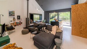 A modern living room with a large gray sectional sofa, a small round coffee table, and a flat-screen TV. There is a fireplace, a dining area with a table and chairs, and a large window offering a view of greenery outside. - Charming Home With Sauna (Paliseul)
