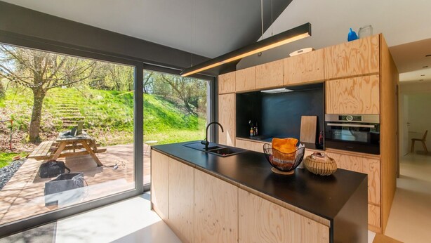 A modern kitchen with wooden cabinetry, a black countertop, and a large window overlooking a garden. - Charming Home With Sauna (Paliseul)