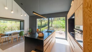 A modern kitchen with a central island, wooden cabinets, and large windows offering a view of greenery. - Charming Home With Sauna (Paliseul)