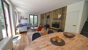 A modern living room with a wooden floor, a grey sofa, a round dining table with wicker baskets, and a fireplace. - Villa in Durbuy With Jacuzzi & Golf Access (Durbuy)