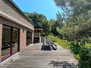 Una terraza de madera con una mesa de picnic y sillas, una parrilla y un vidrio corredizo que da a un patio.