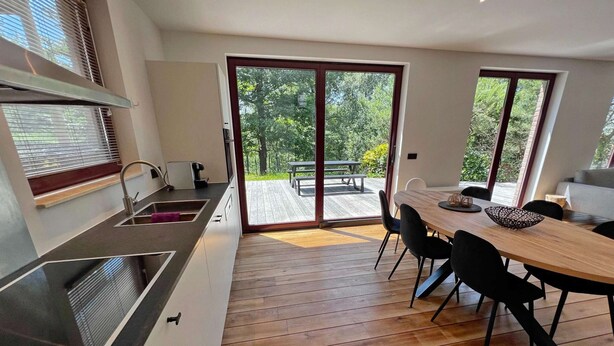 A modern kitchen with a large island, a dining area with a wooden table and chairs, and a sliding glass door leading to an outdoor deck. - Villa in Durbuy With Jacuzzi & Golf Access (Durbuy)
