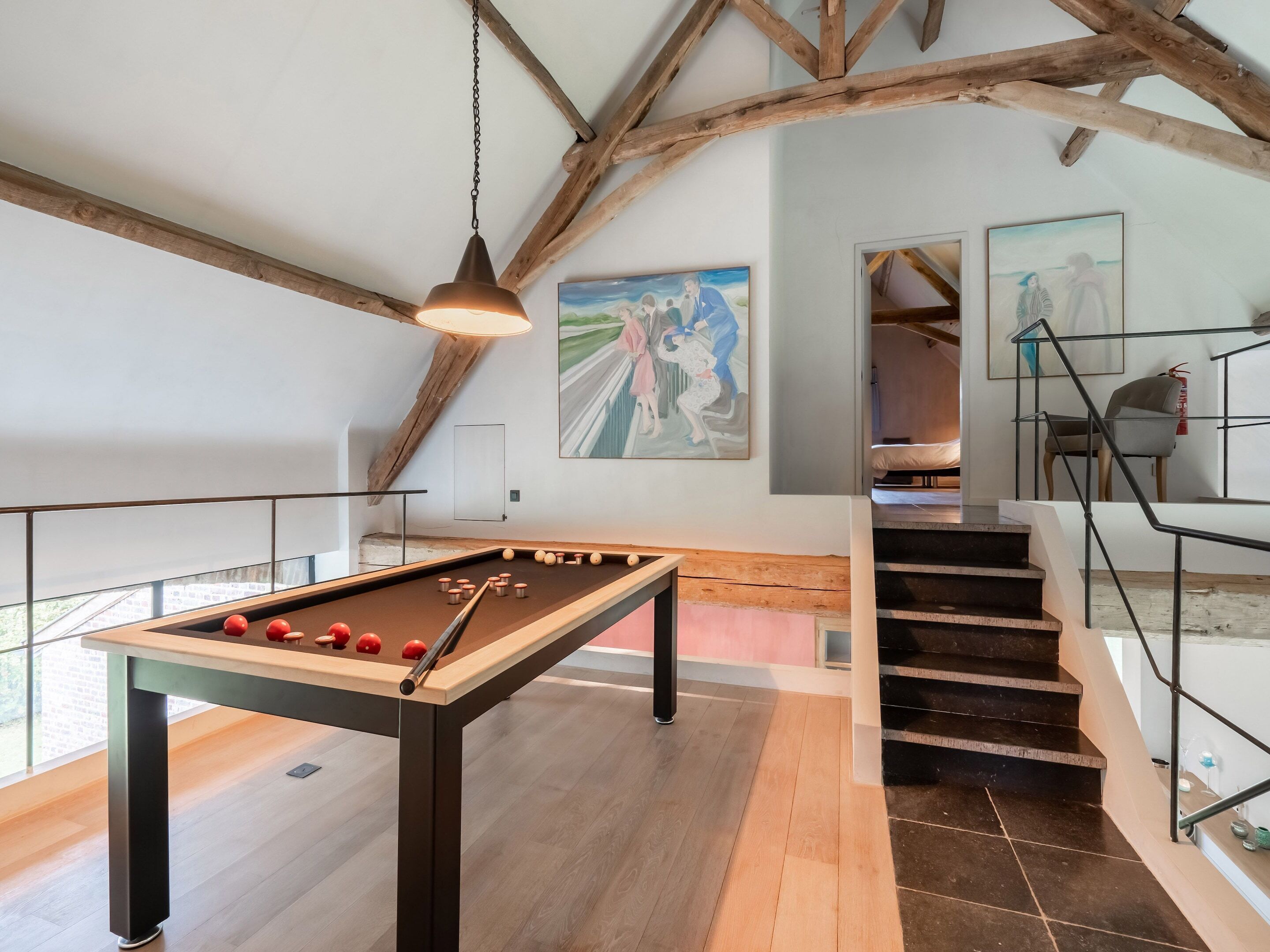 A pool table in a room with exposed wooden beams, a staircase, and artwork on the walls.
