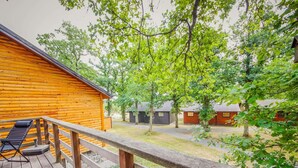 A wooden deck with a lounge chair and a wooden railing overlooking a wooded area with cabins. - Chalet Le Beau Renard, Heure (Somme-Leuze)