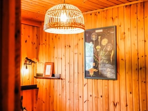 A wooden interior with a hanging light fixture, a framed artwork of flowers, and a small shelf with a framed item. - Chalet Le Beau Renard, Heure (Somme-Leuze)