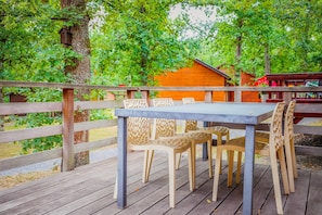A wooden deck with a table and chairs, surrounded by trees and a wooden cabin in the background. - Chalet Le Beau Renard, Heure (Somme-Leuze)