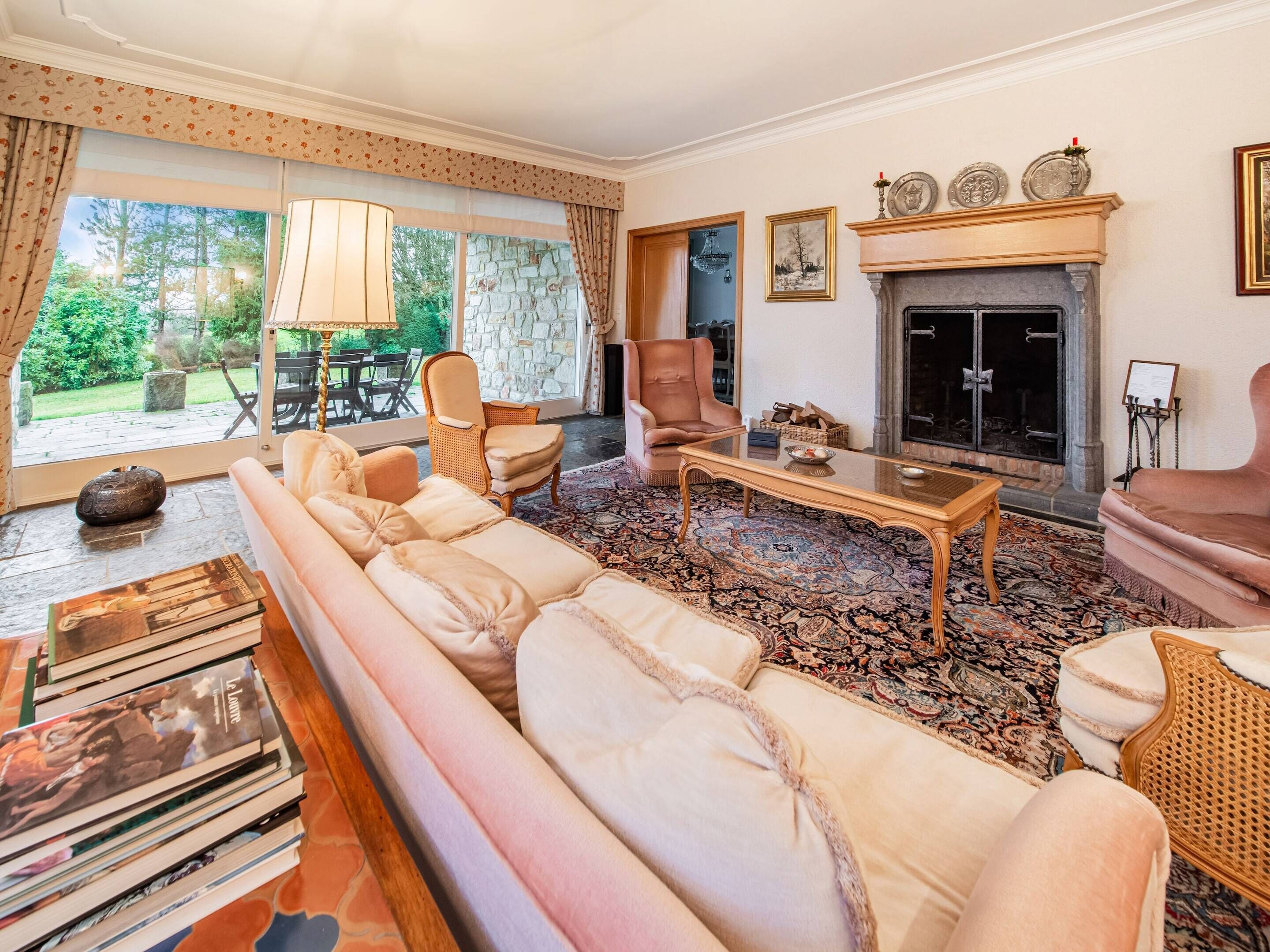 A spacious living room with a large sofa, two armchairs, a coffee table, and a fireplace. There is a stone wall and a large window with curtains.