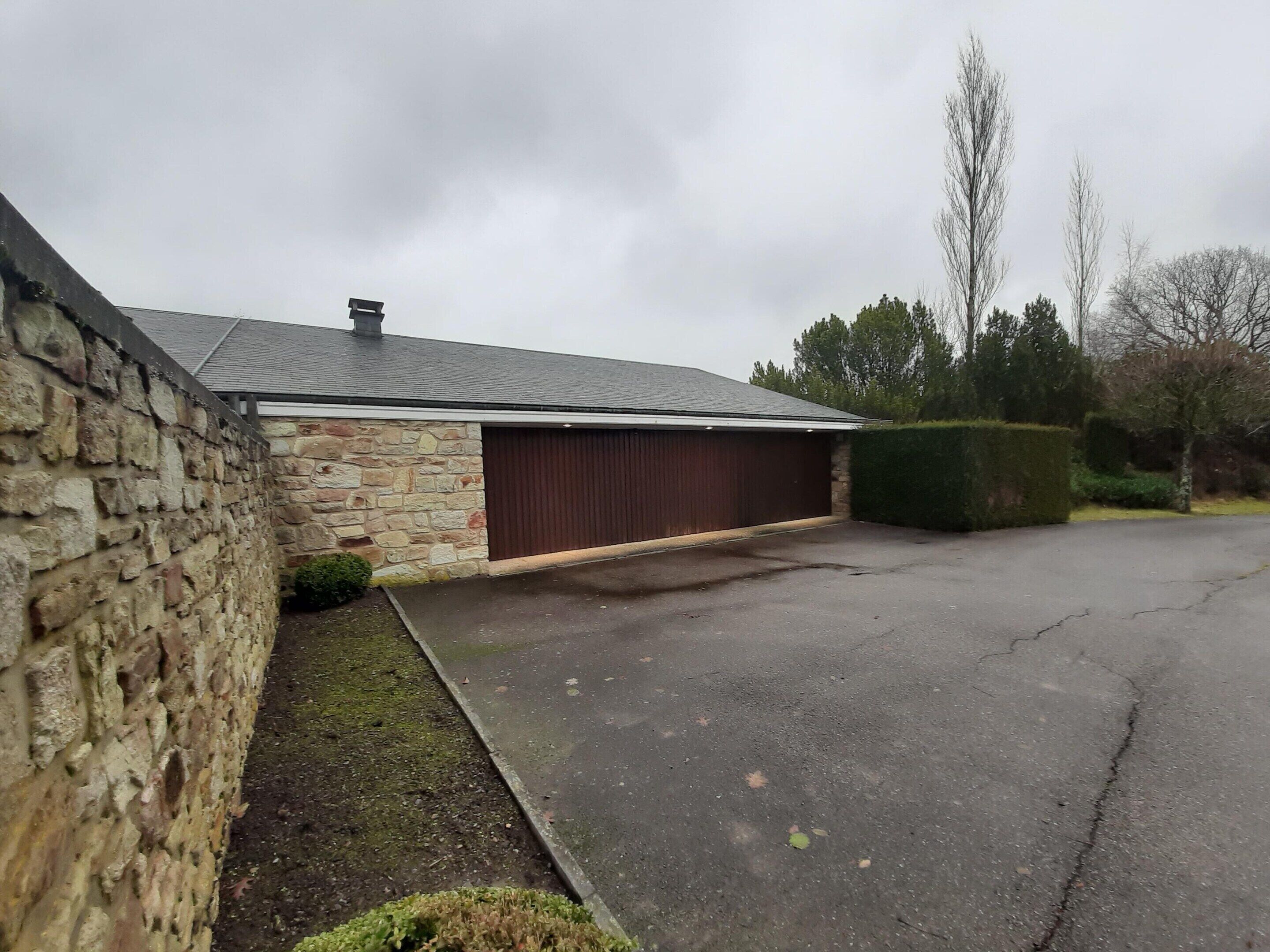 A stone building with a garage, a paved driveway, and a hedge.