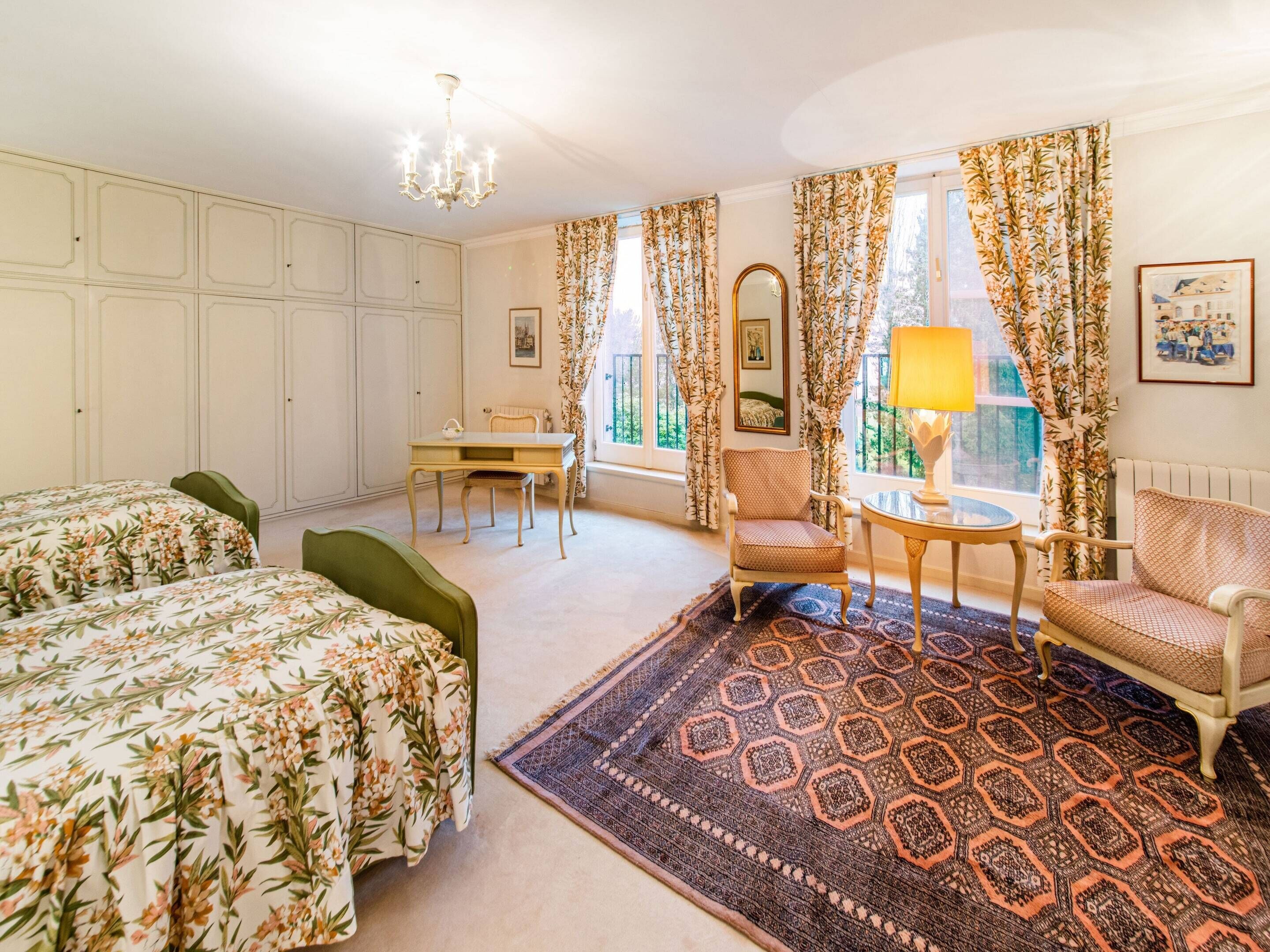 A room with a bed, two chairs, a table, a lamp, a mirror, and a patterned rug.