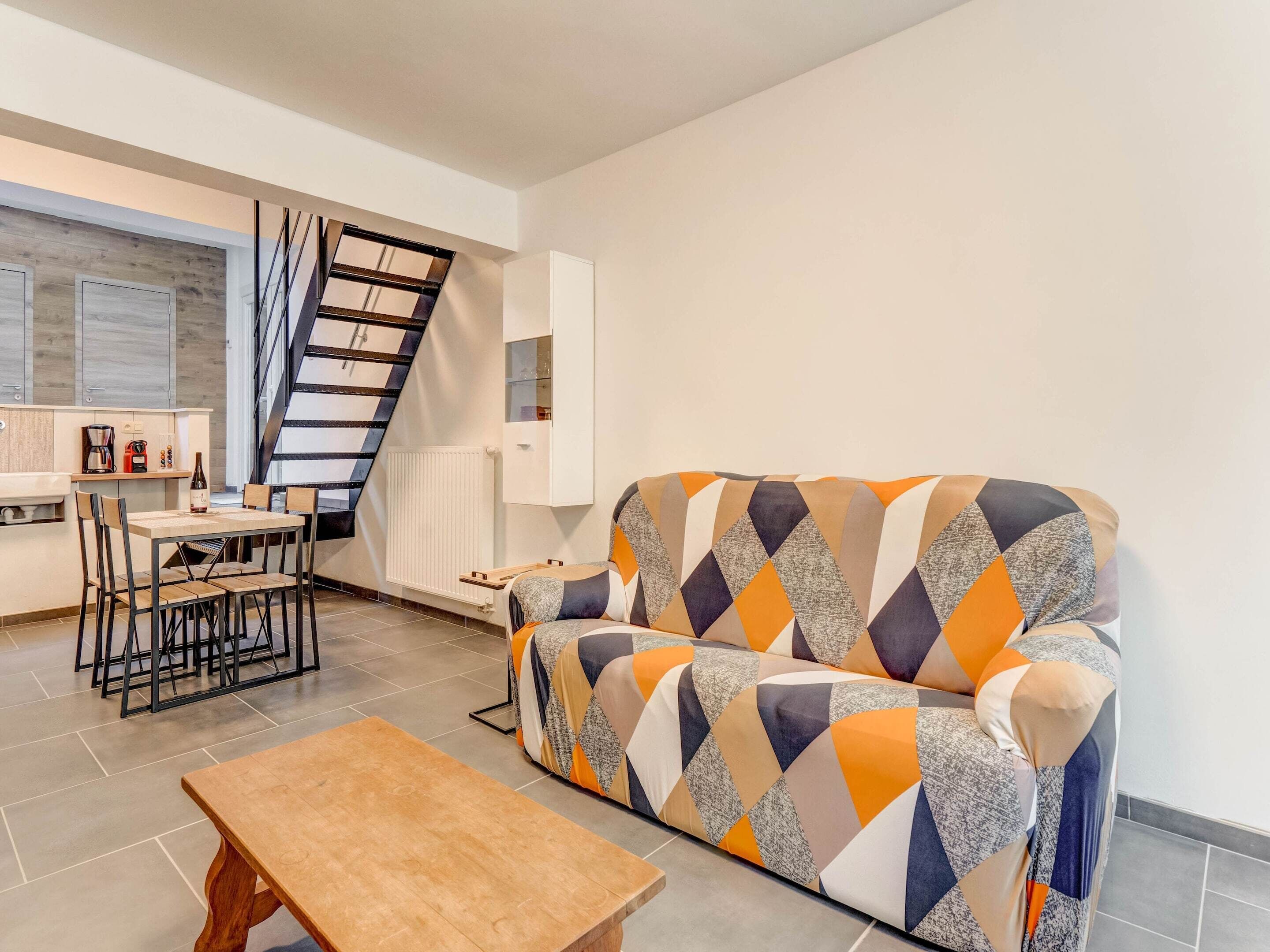 Un salon moderne comprenant un canapé, une table basse en bois, un coin repas avec des chaises, une cuisine avec un comptoir et des appareils électroménagers, et un escalier.