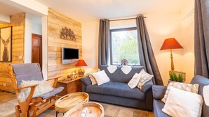 A cozy living room with a wooden wall, a grey sofa, a wooden rocking chair, a TV, and a window with curtains. - Farmhouse Kalys in Vielsalm With 180° View of the Forest (Vielsalm)