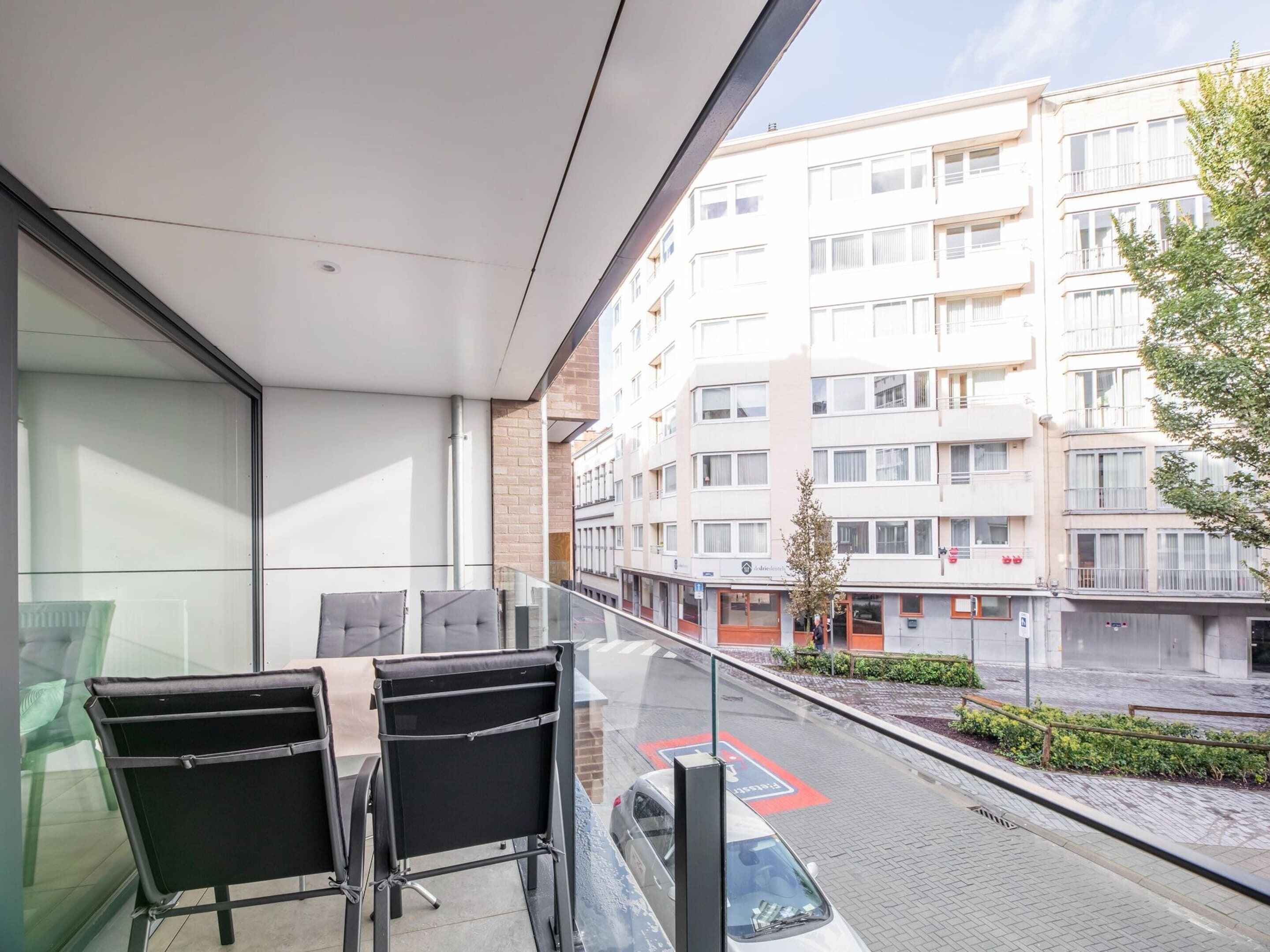 A balcony with two black chairs and a table, overlooking a street with a parked car and a building with multiple windows.