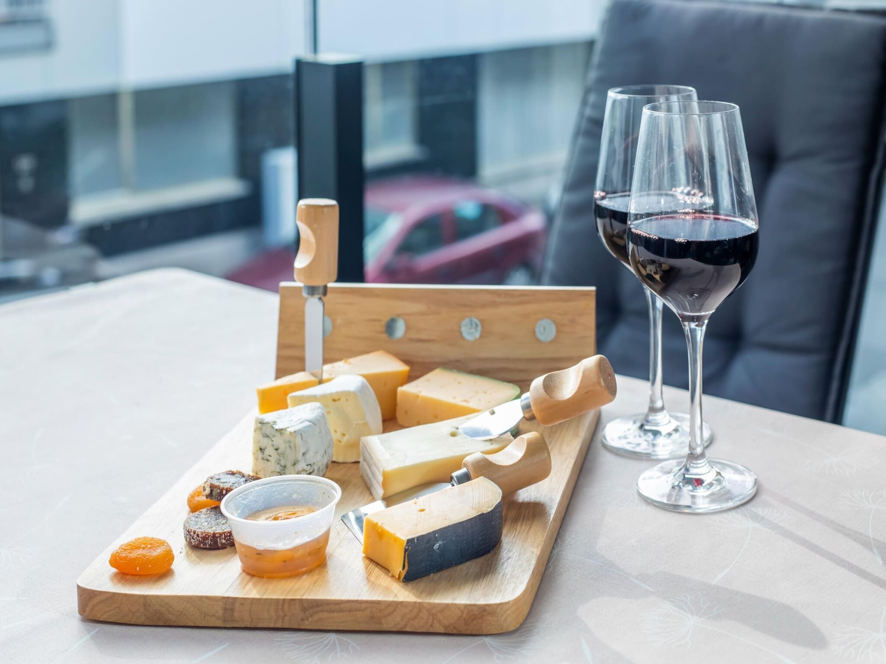 Una tabla de quesos con distintos tipos de queso y acompañamientos, dos copas de vino tinto, y un cuchillo sobre una mesa con vistas a un paisaje urbano.