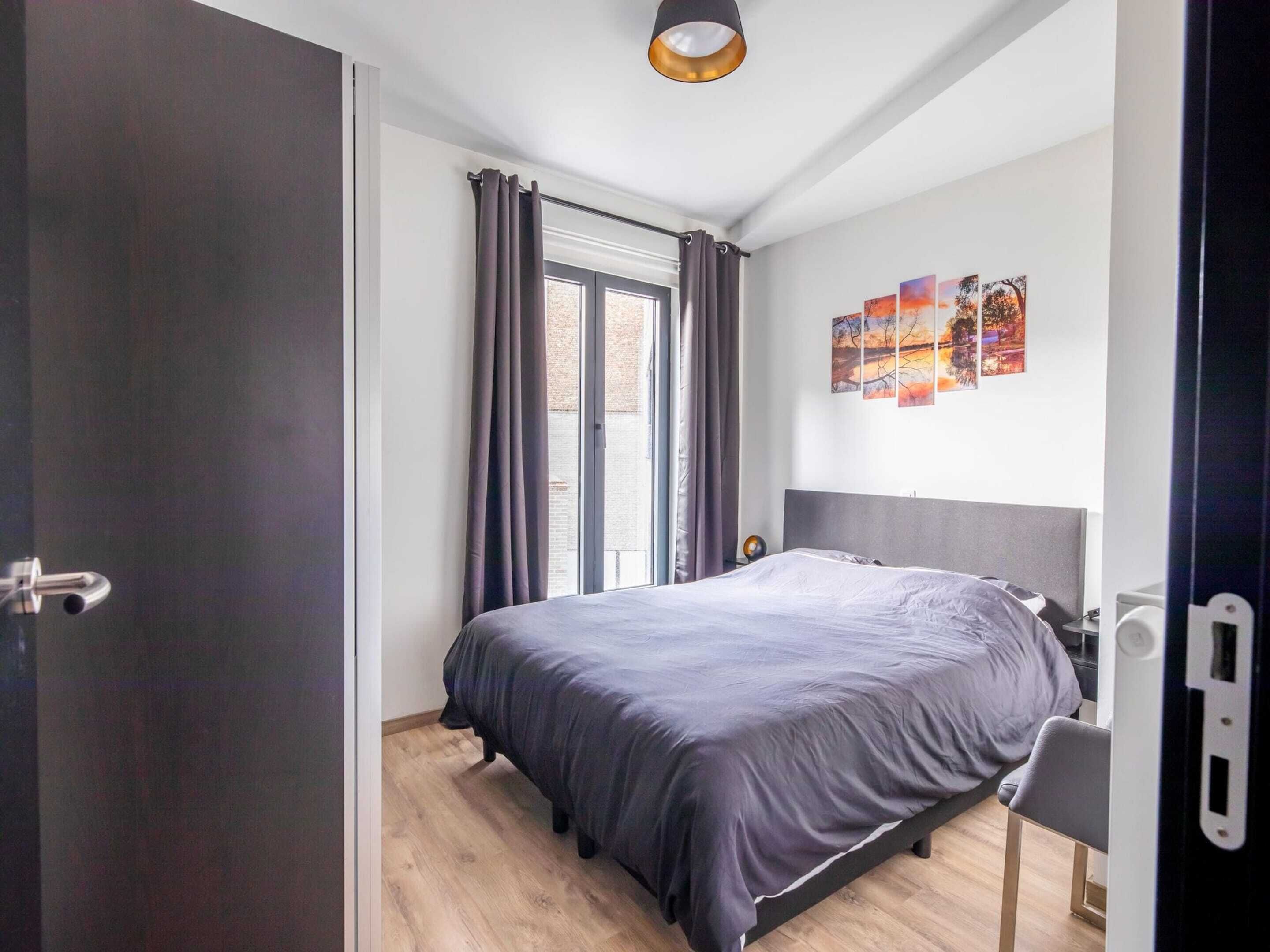 A modern bedroom with a large bed, a window with curtains, a bedside table, and a wall with artwork.