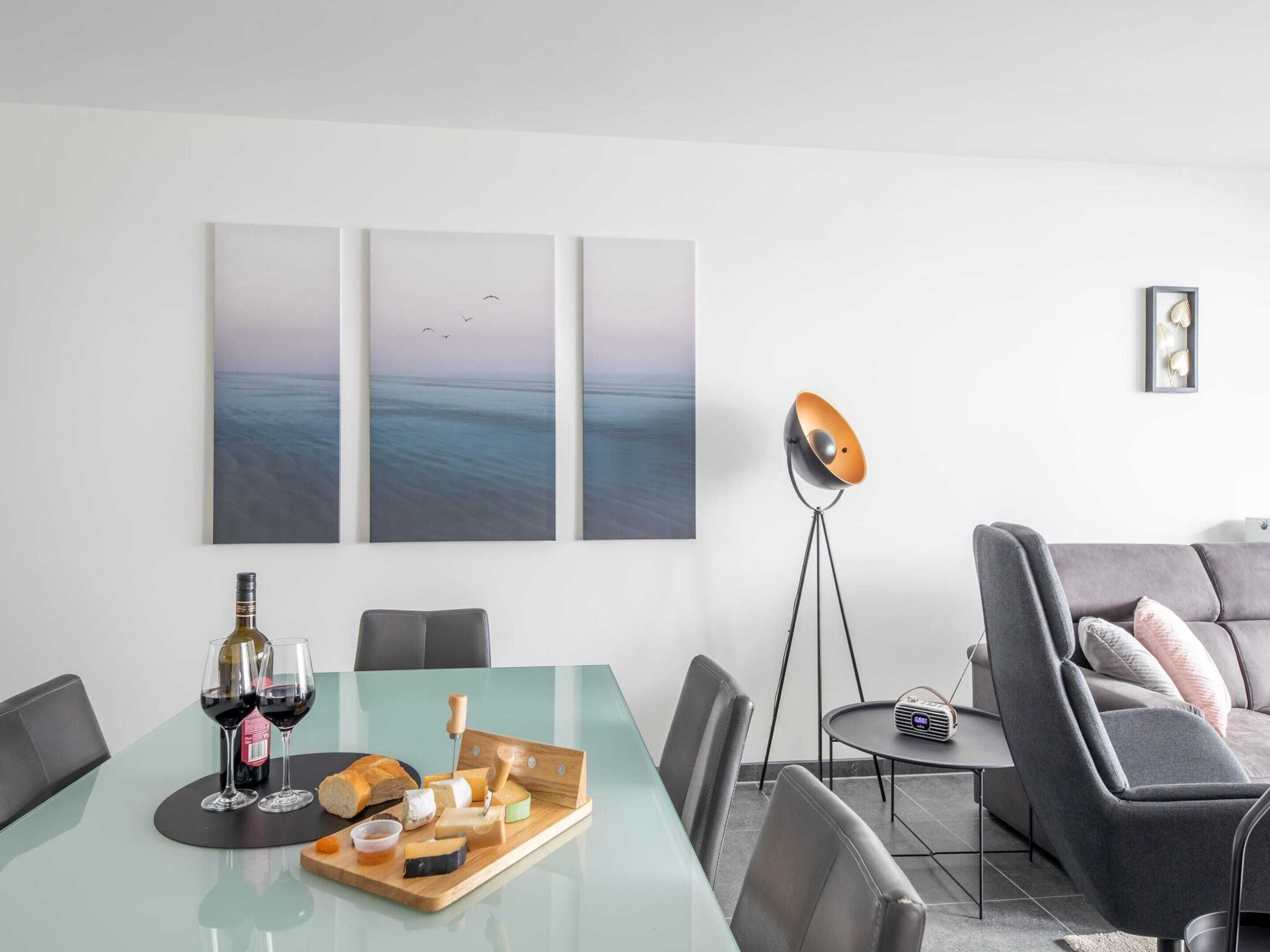 A modern living room with a glass dining table set for a meal, a grey sofa, a floor lamp, and three large wall art pieces depicting a seascape.