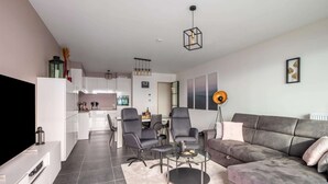 A modern living room with a grey sofa, a coffee table, and a TV. There is a kitchen in the background with white cabinets and a dining area with chairs. - Cosy Apartment in Oostende With Private Terrace (Ostend)
