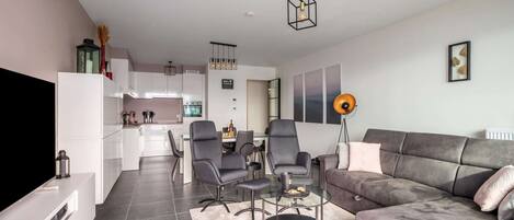 A modern living room with a grey sofa, a coffee table, and a TV. There is a kitchen in the background with white cabinets and a dining area with chairs.