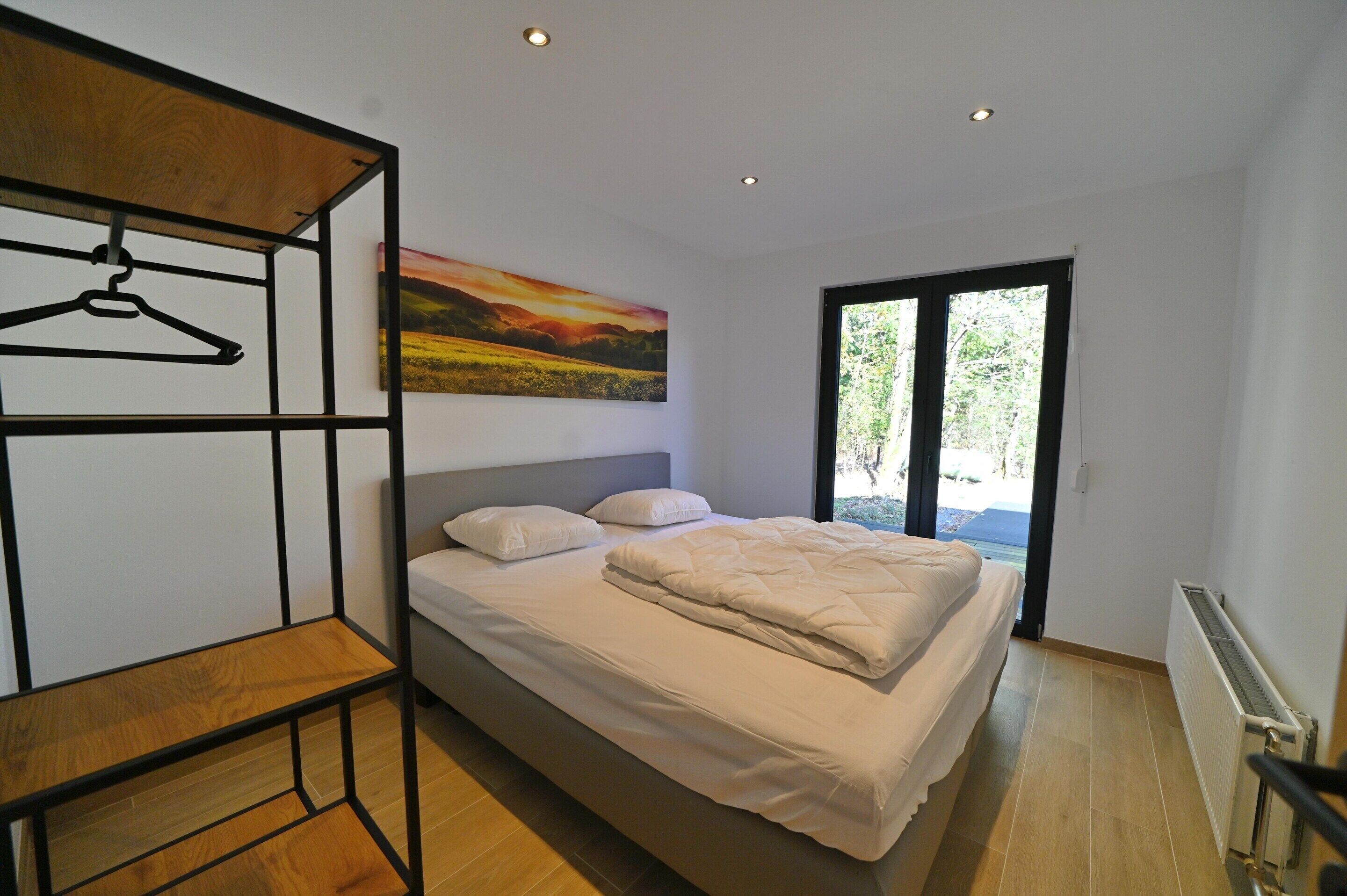 A bedroom with a bed, a wooden shelf, a painting, and a view through a window.