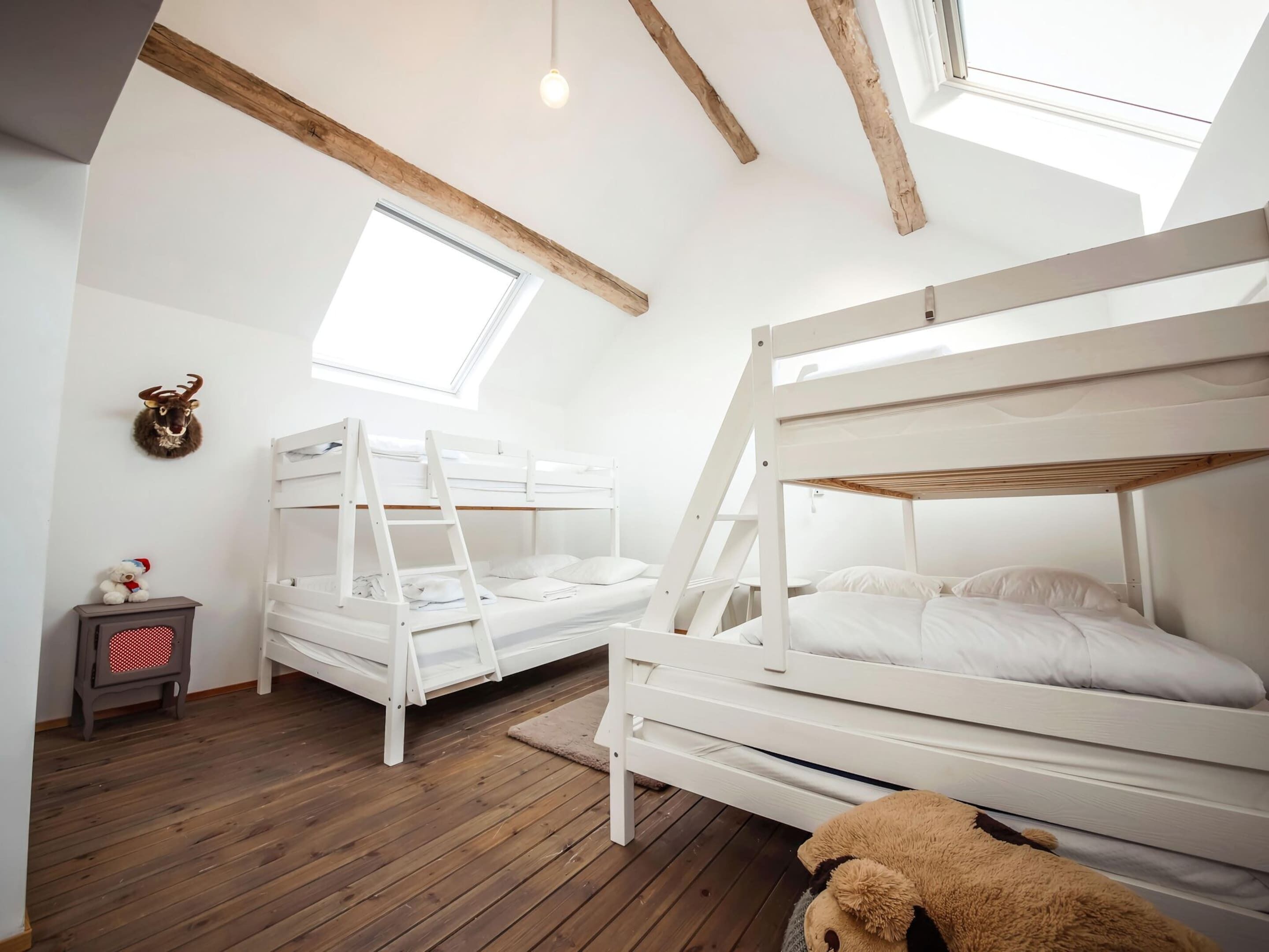 A room with two bunk beds, wooden beams on the ceiling, a small table with a teddy bear, and a mounted deer head.