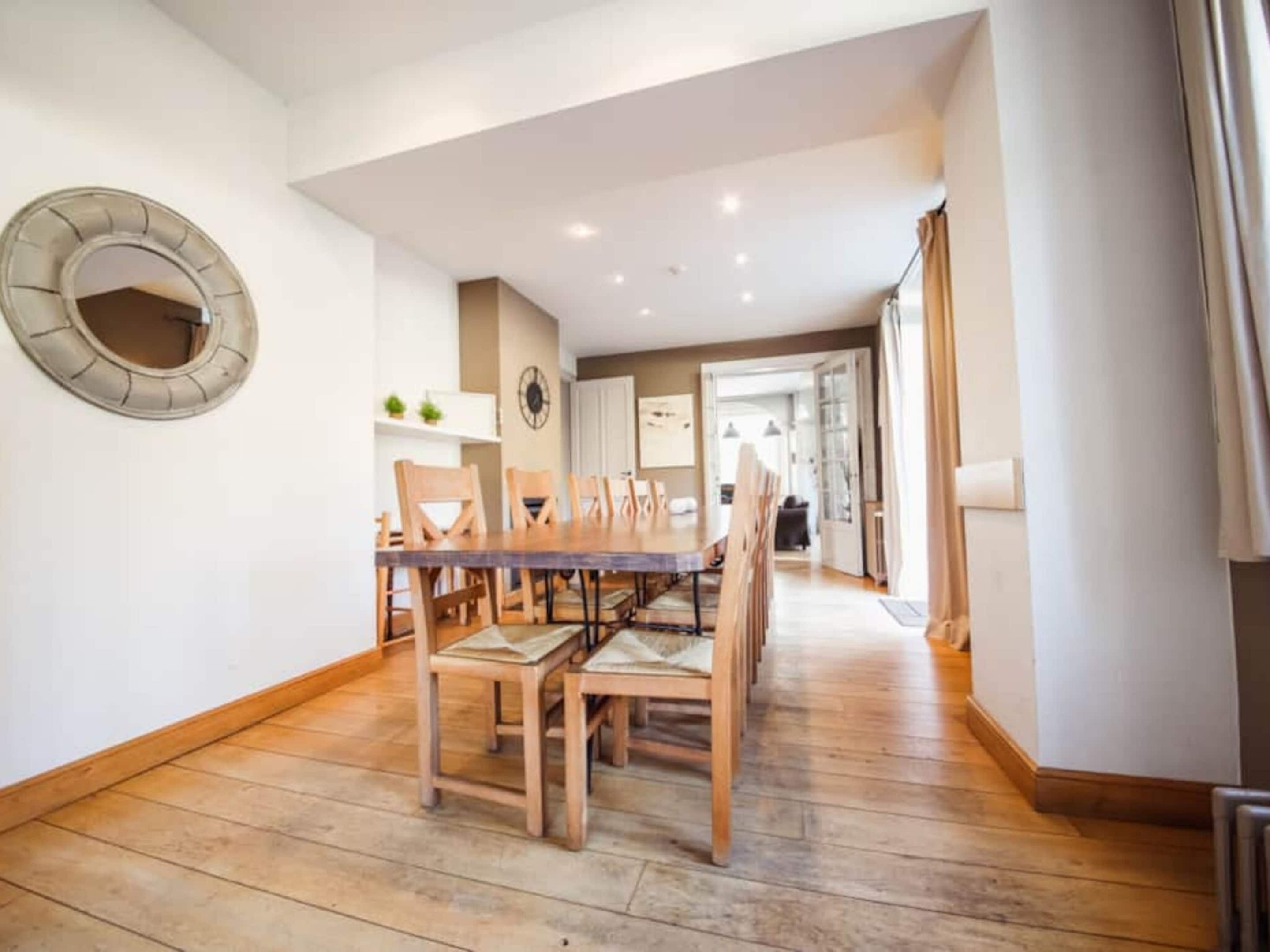 Un coin repas avec une table et des chaises en bois, un miroir rond accroché au mur et une cuisine en arrière-plan.
