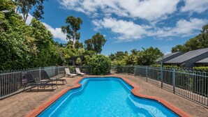 A rectangular swimming pool with a brick border, surrounded by a metal fence, and shaded by trees. - Sunset Waters 19 Hamilton Island - golf buggy and transfers (Hamilton Island)