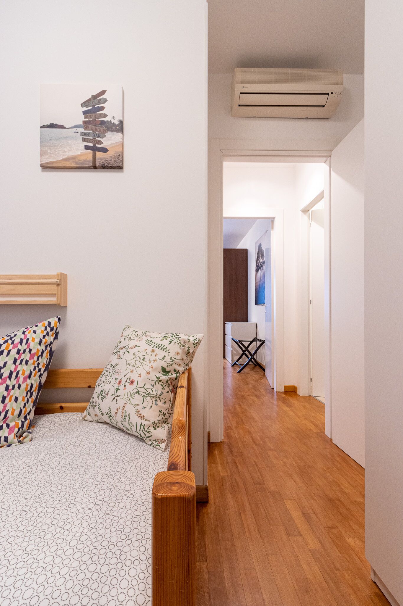 Une chambre à coucher avec un lit en bois, un coussin à motifs, un climatiseur fixé au mur et un tableau représentant une scène de plage.