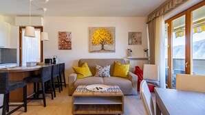 A modern living room with a sofa, coffee table, and dining area. There is a painting of a tree on the wall and a window with curtains. - Wrap around Lake Views Family sized condo with large balcony (Campione d'Italia)