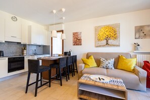 A modern kitchen with white cabinets, a dining area with bar stools, a sofa with yellow cushions, and a coffee table.