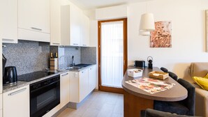 A compact kitchen with a dishwasher, stove, and a coffee maker. There's a dining area with a table, chairs, and a lamp. - Wrap around Lake Views Family sized condo with large balcony (Campione d'Italia)